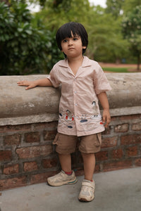 A young boy wearing a light pink short-sleeve button-up shirt with cartoon graphics, brown shorts, and white sneakers, leaning against a concrete pillar in an outdoor park setting.