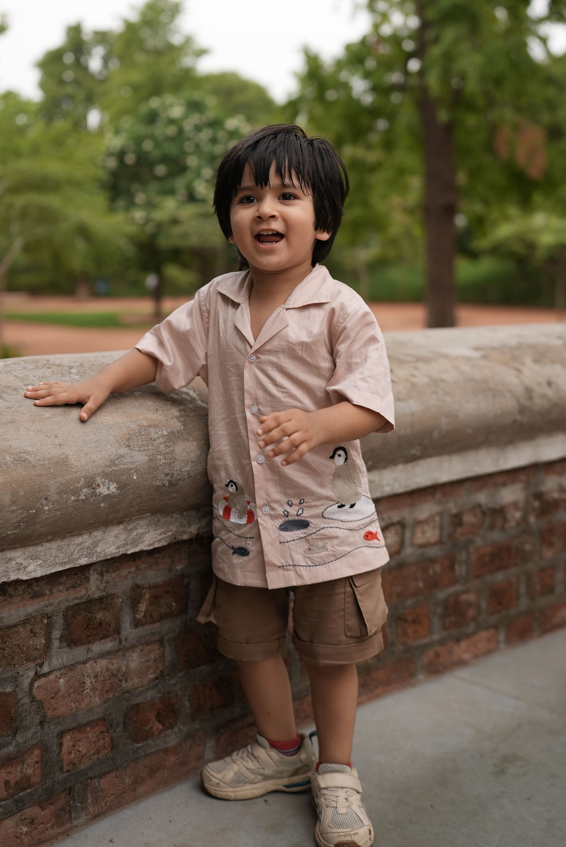 A young boy wearing a light pink patterned short-sleeve shirt, brown shorts, and white sneakers, leaning against a concrete wall outdoors with trees in the background.