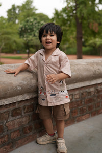 A young boy wearing a light pink patterned short-sleeve shirt, brown shorts, and white sneakers, leaning against a concrete wall outdoors with trees in the background.