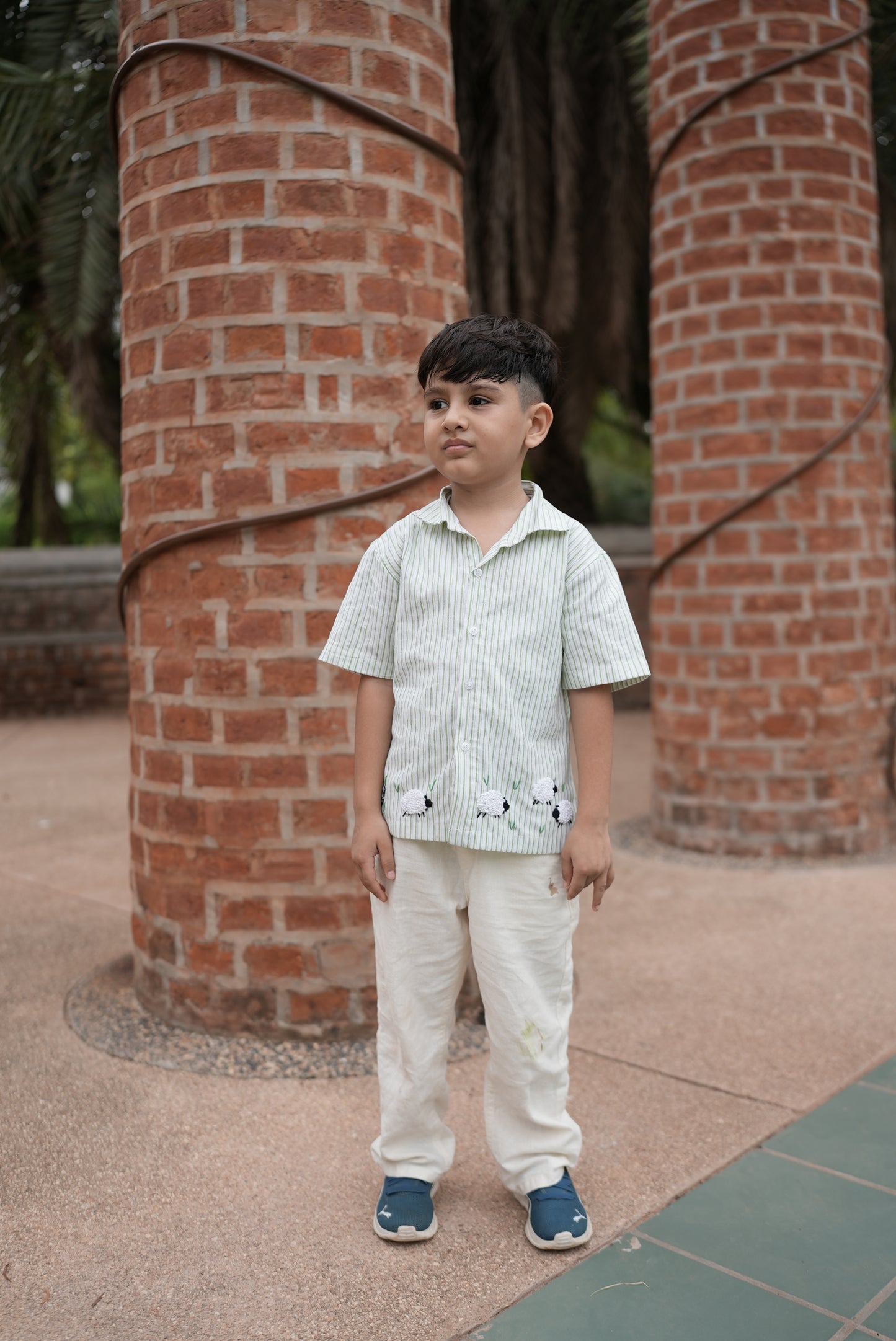 A young boy stands outdoors in front of two large red brick columns, wearing a light green and white striped short-sleeve shirt with black spider graphics on the front, cream trousers, and blue sneakers.