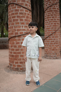 A young boy stands outdoors in front of two large red brick columns, wearing a light green and white striped short-sleeve shirt with black spider graphics on the front, cream trousers, and blue sneakers.