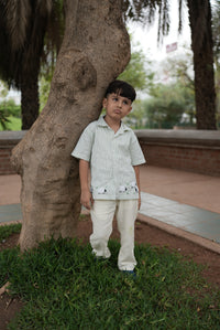 A young boy stands outdoors leaning against a large tree trunk in a park-like setting. He is wearing a light green and white vertically striped short-sleeve button-up shirt with small embroidered details near the hem, paired with off-white trousers.