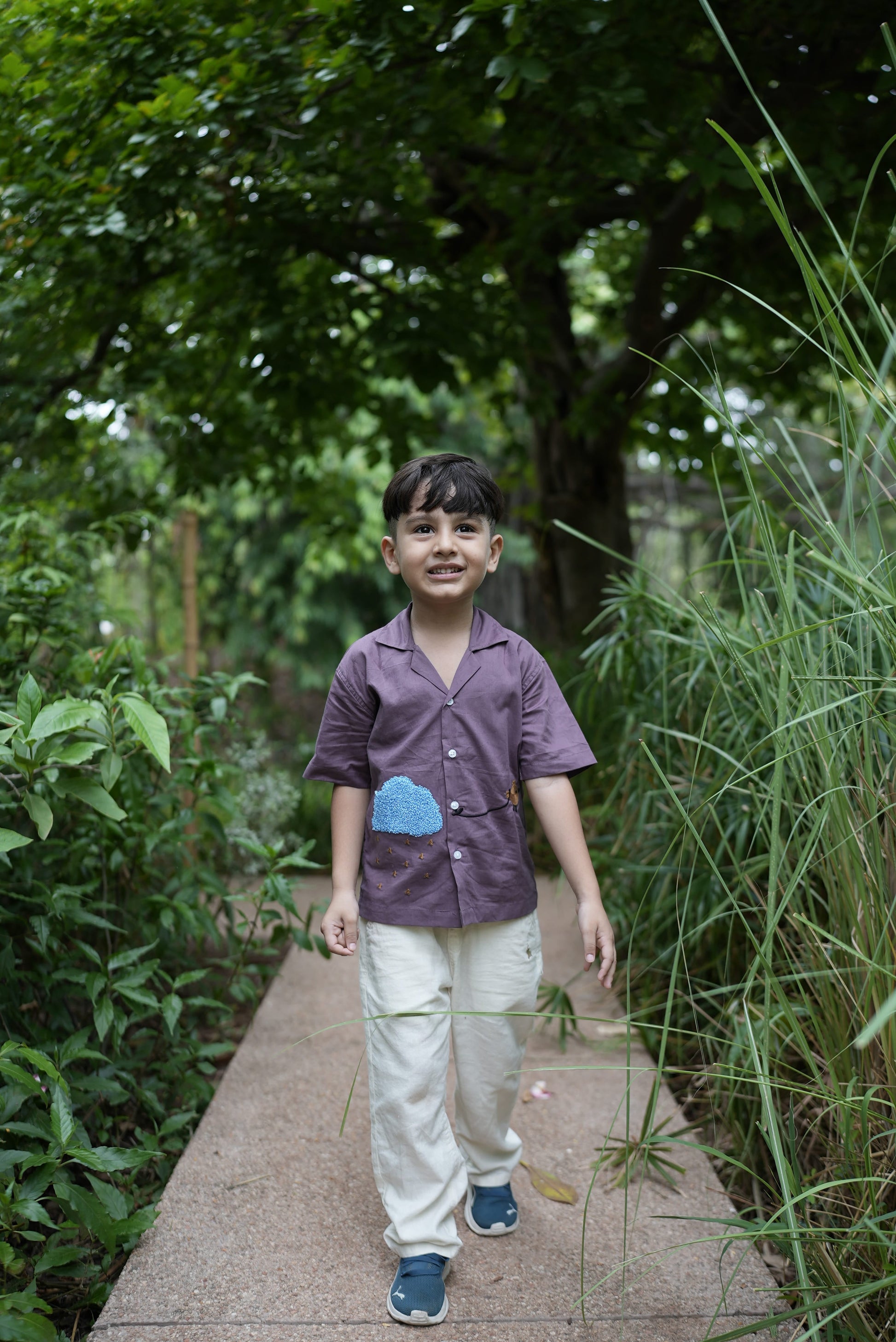 A young boy standing on a paved path outdoors, wearing a purple short-sleeve button-up shirt with a light blue cloud patch on the chest, paired with off-white pants and blue sneakers. The background is filled with lush green foliage and tall grass.