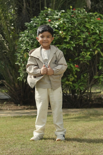 Child in a beige jacket and pants standing outdoors with greenery in the background
