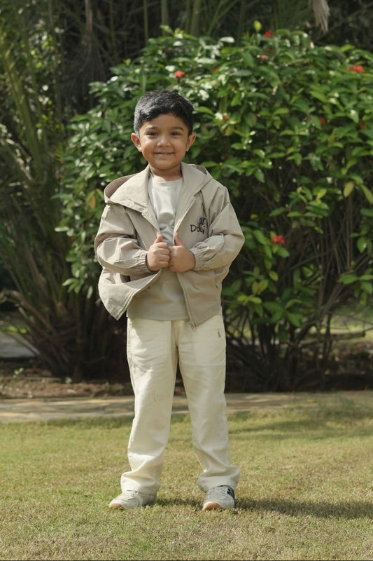 Child in a beige jacket and pants standing outdoors with greenery in the background