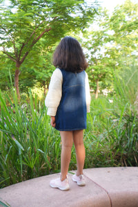 A child viewed from behind wearing a white long-sleeve top, a dark blue denim vest, and white sneakers, standing on a paved path outdoors with tall green grass and trees in the background.