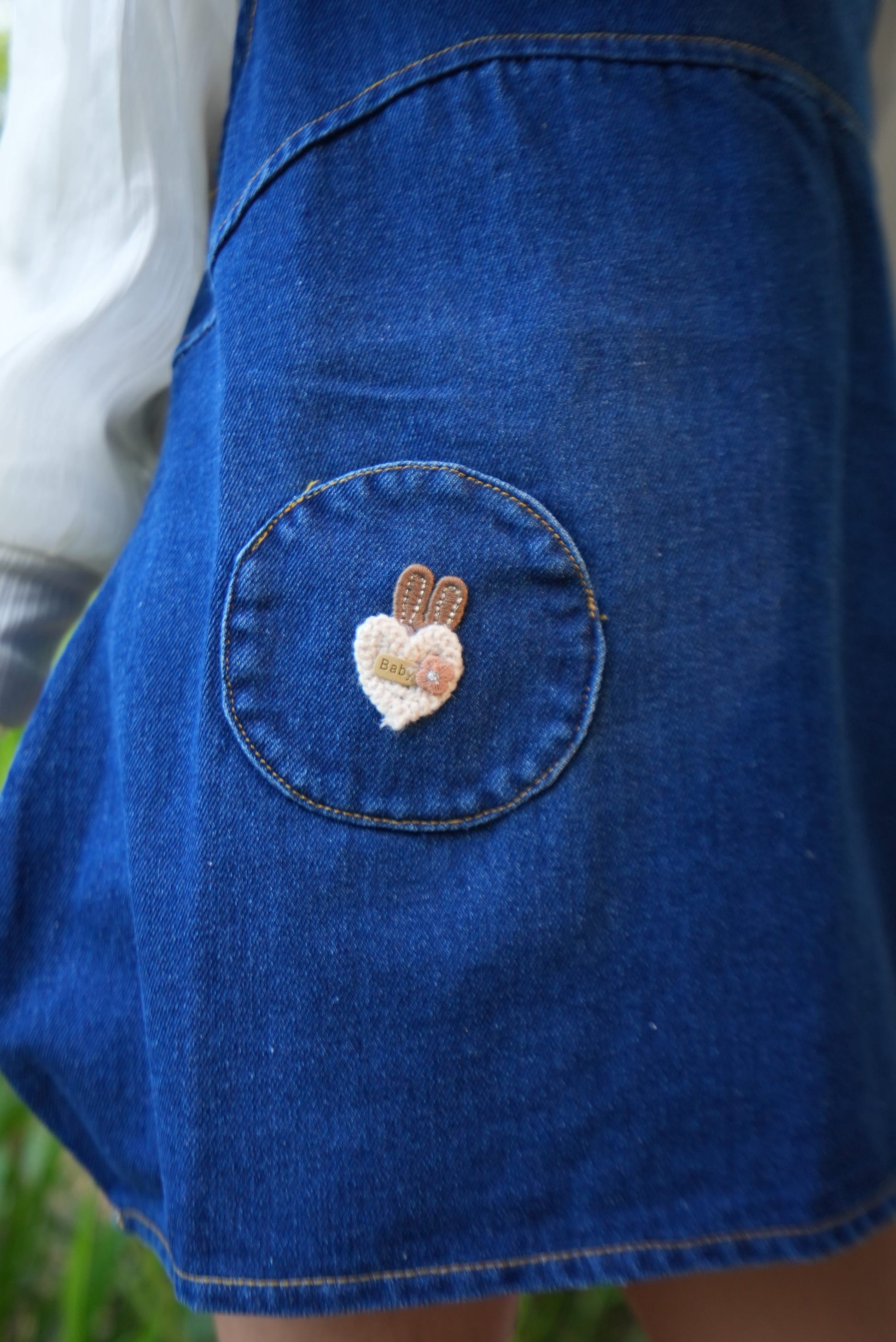 A close-up of a blue denim garment, likely a skirt or shorts, featuring a circular patch pocket with a small embroidered bunny detail in cream and brown.