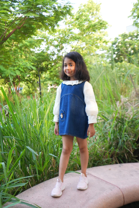 A young girl wearing a blue denim pinafore dress over a white long-sleeve shirt and white sneakers, standing outdoors on a paved path surrounded by tall green grass and trees.