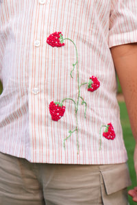 A close-up of a person wearing a short-sleeve button-up shirt with thin red and white vertical stripes and embroidered red strawberries with green stems on the front. The person is also wearing khaki cargo pants, with a blurred green outdoor background.
