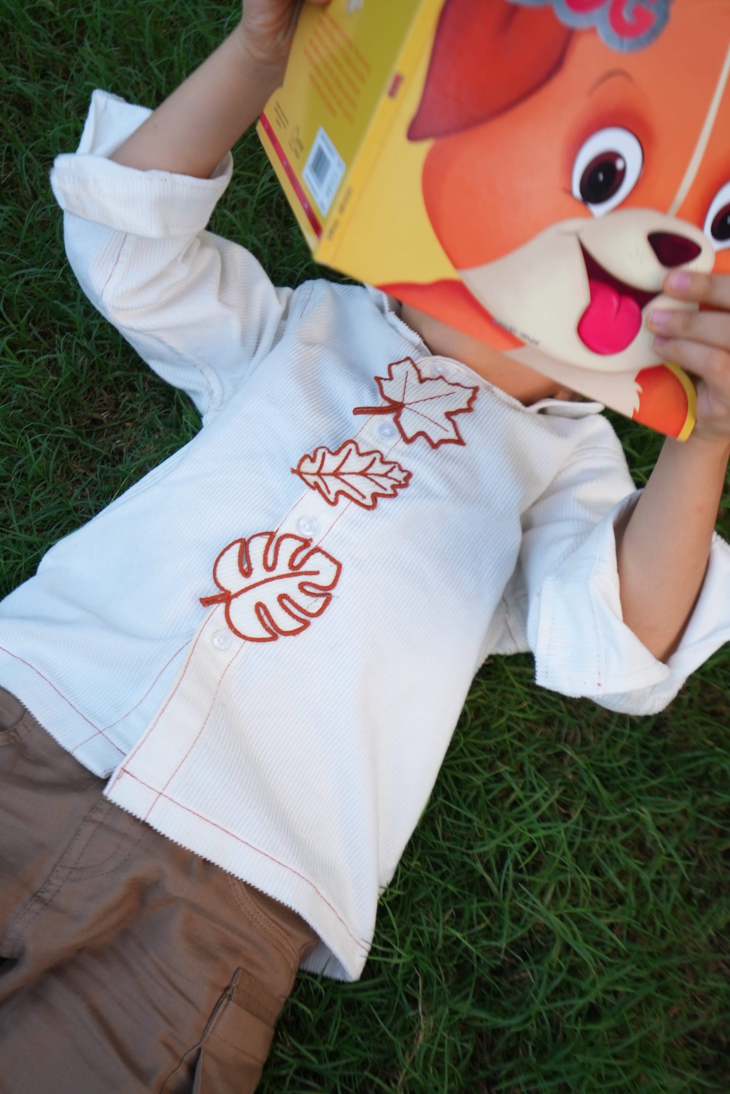A child lying on green grass wearing a white long-sleeve shirt with red embroidered leaf motifs. The child is holding a yellow box with a cartoon dog face.