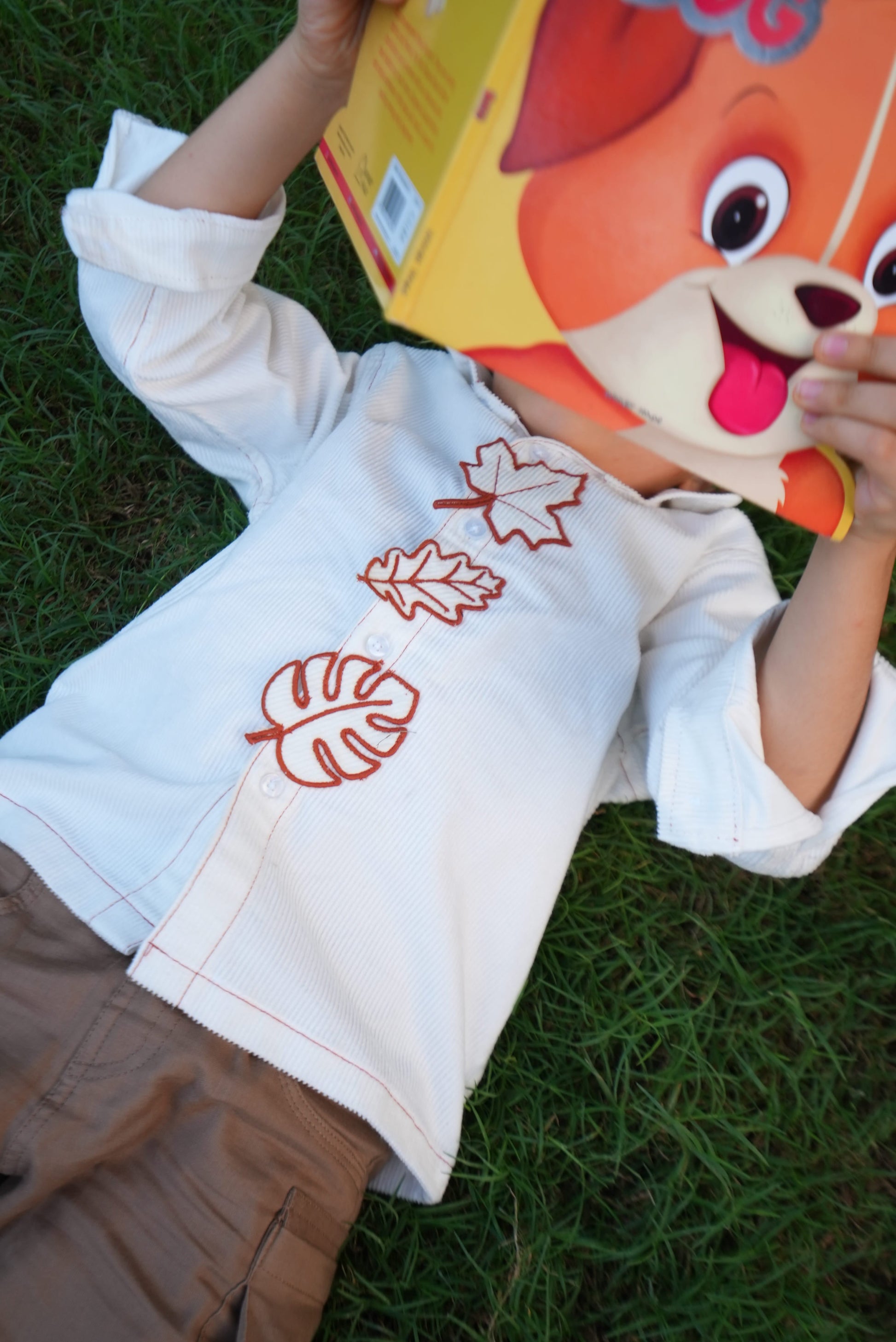 A child lying on green grass wearing a white long-sleeve shirt with red embroidered leaf motifs. The child is holding a yellow box with a cartoon dog face.