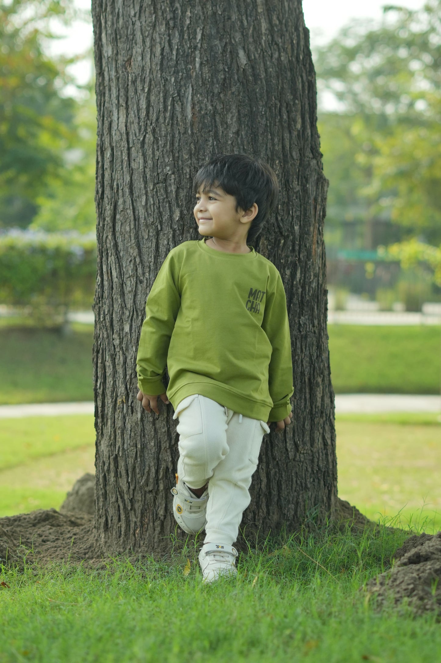 Person wearing a green jacket and white pants standing against a tree in a park.