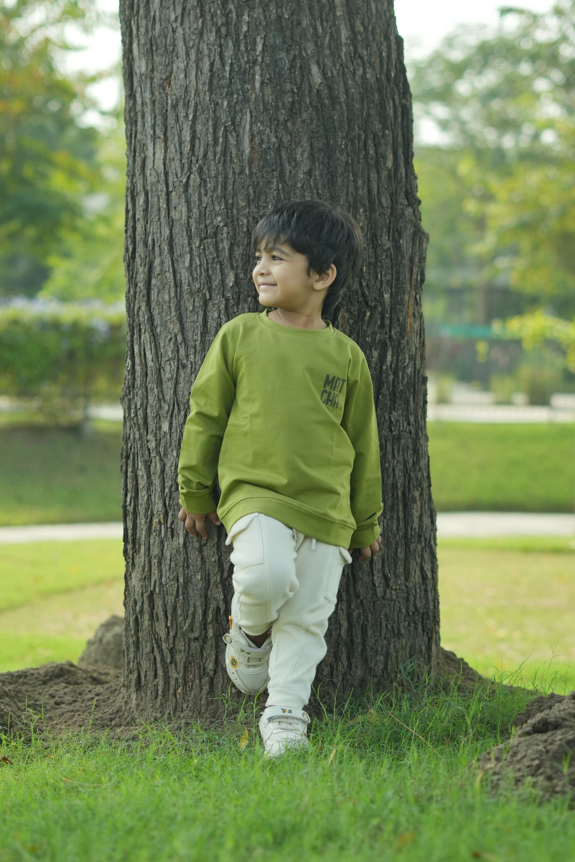 Person wearing a green jacket and white pants standing against a tree in a park.