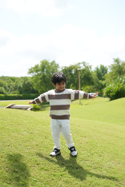 A young boy stands on a grassy lawn with his arms outstretched, wearing a brown and cream striped knit sweater, white pants, and black-and-white sneakers. Trees and a bright sky are visible in the background.