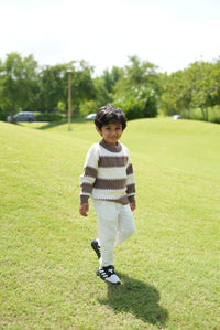 A young boy walking on a grassy park hill wearing a brown and cream striped knit sweater, white pants, and black and white sneakers.