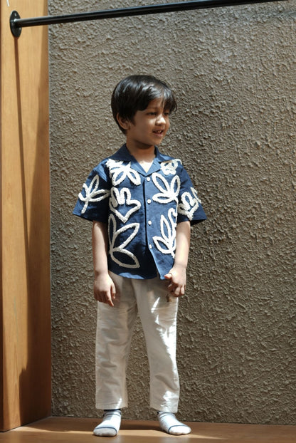 Child wearing a blue floral shirt and white pants standing against a textured wall.