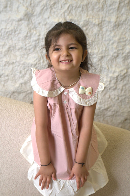Young girl wearing a pink dress with white ruffled collar and cuffs, sitting on a beige couch.