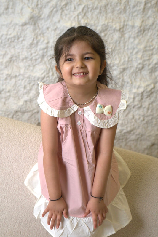 Young girl wearing a pink dress with white ruffled collar and cuffs, sitting on a beige couch.