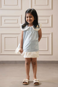Young girl in a light blue dress standing against a white paneled wall.