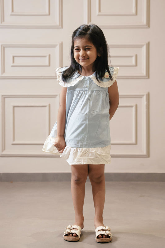 Young girl in a light blue dress standing against a white paneled wall.