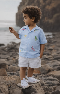 Child wearing a light blue shirt with floral patterns and white shorts standing on rocks by the water.