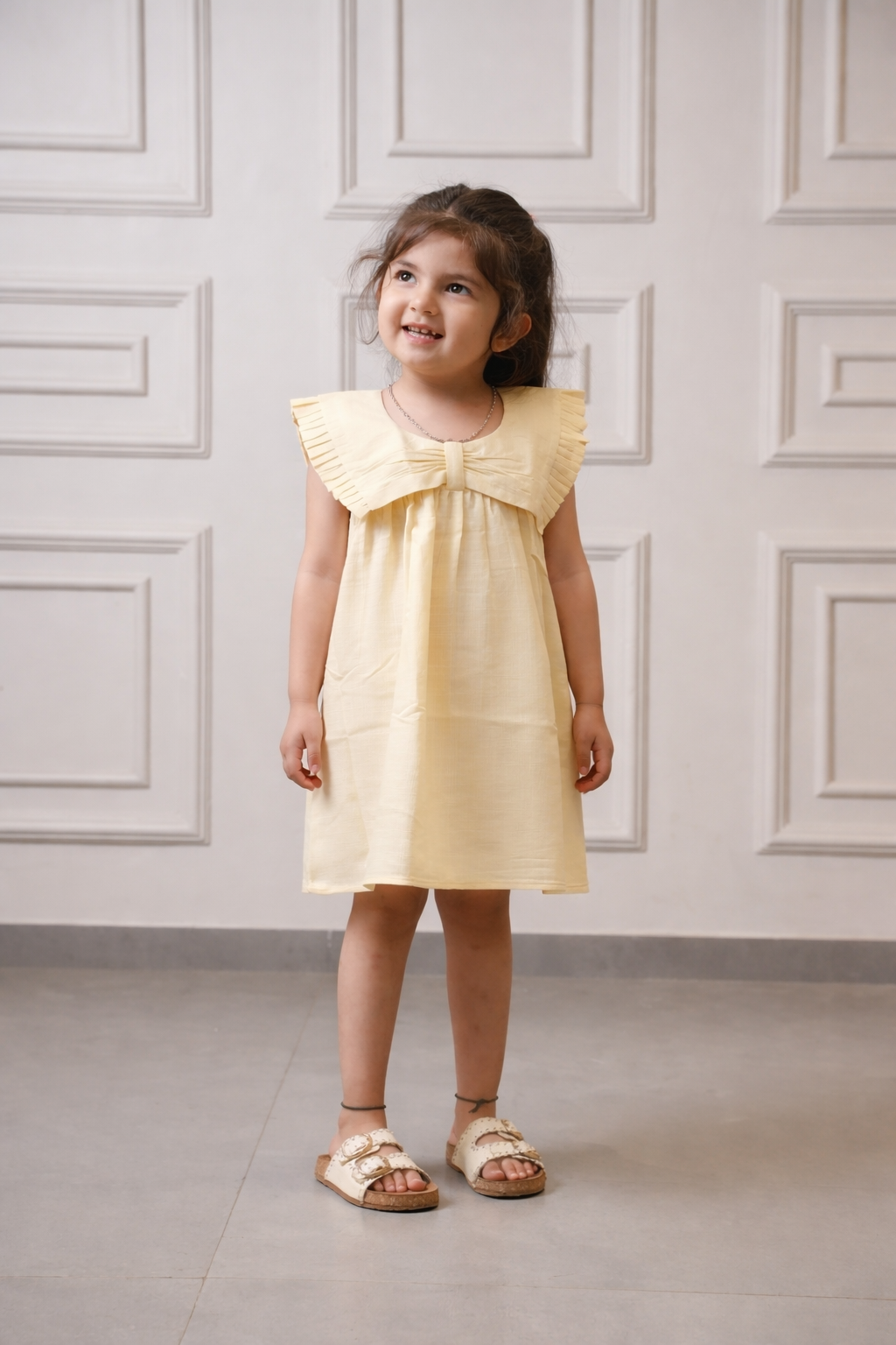 Young girl wearing a yellow dress standing against a white paneled wall.