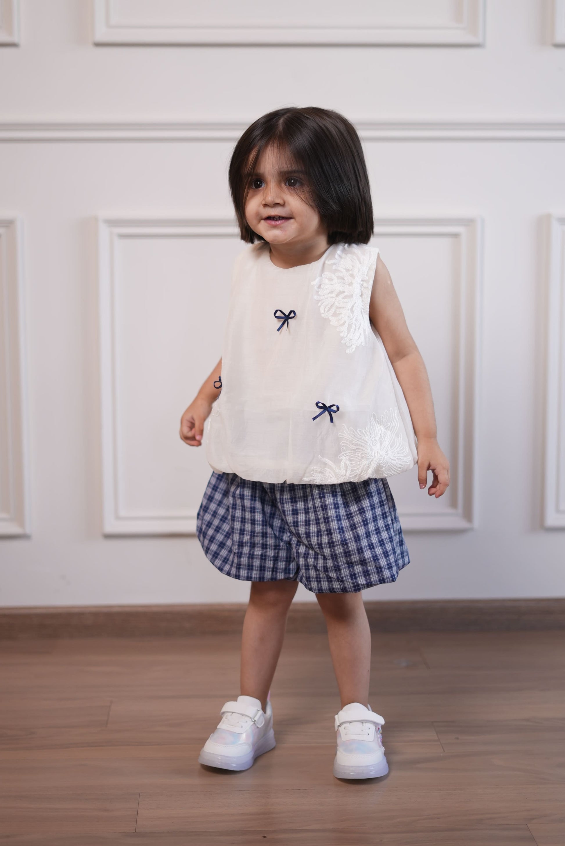 Child wearing a white dress with blue plaid skirt and white shoes on a wooden floor.