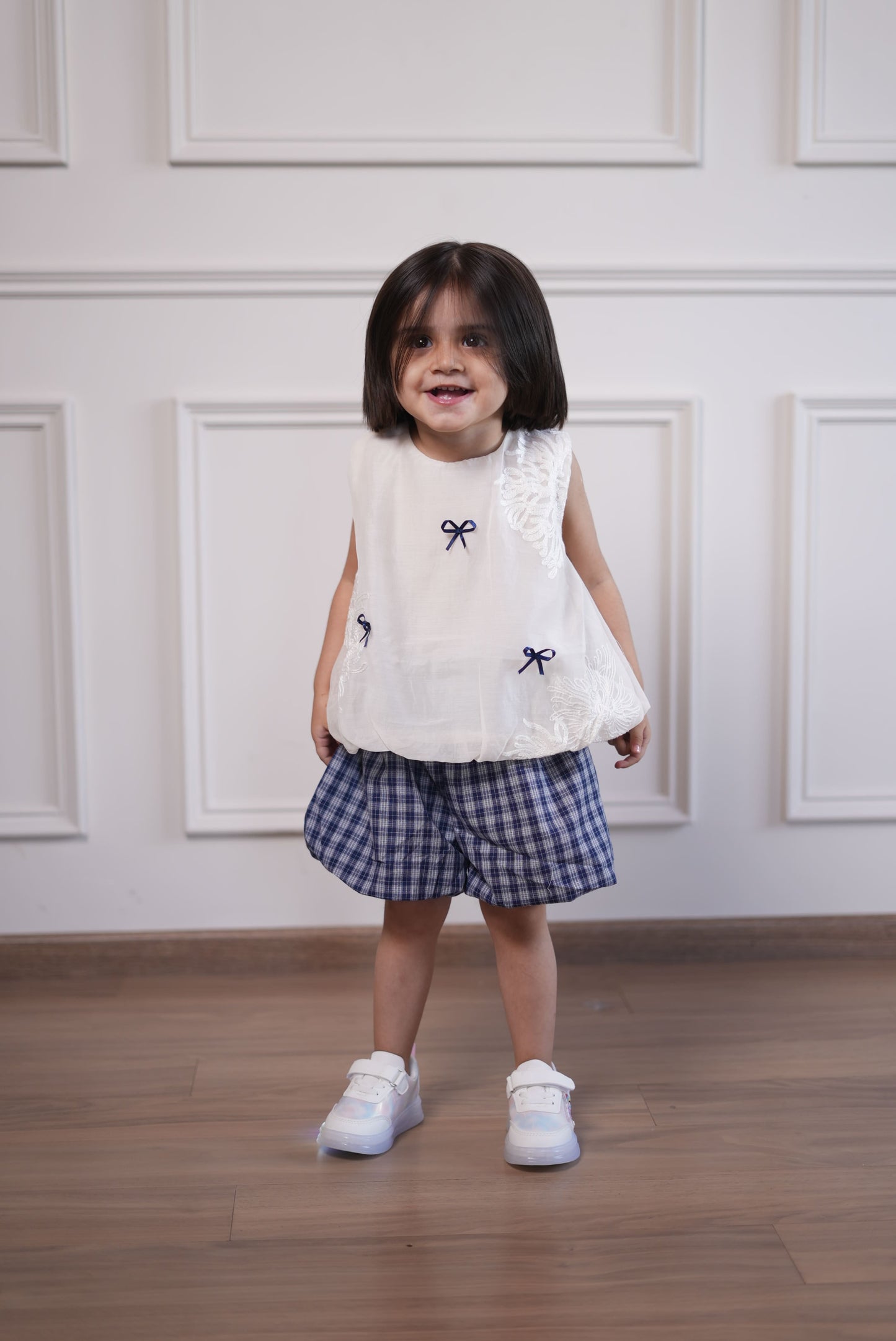 Child wearing a white lace top and plaid skirt with black bow accents on a wooden floor.
