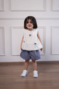 Child wearing a white lace top and plaid skirt with black bow accents on a wooden floor.