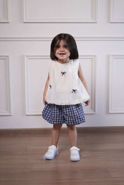 Child wearing a white lace top and plaid skirt with black bow accents on a wooden floor.