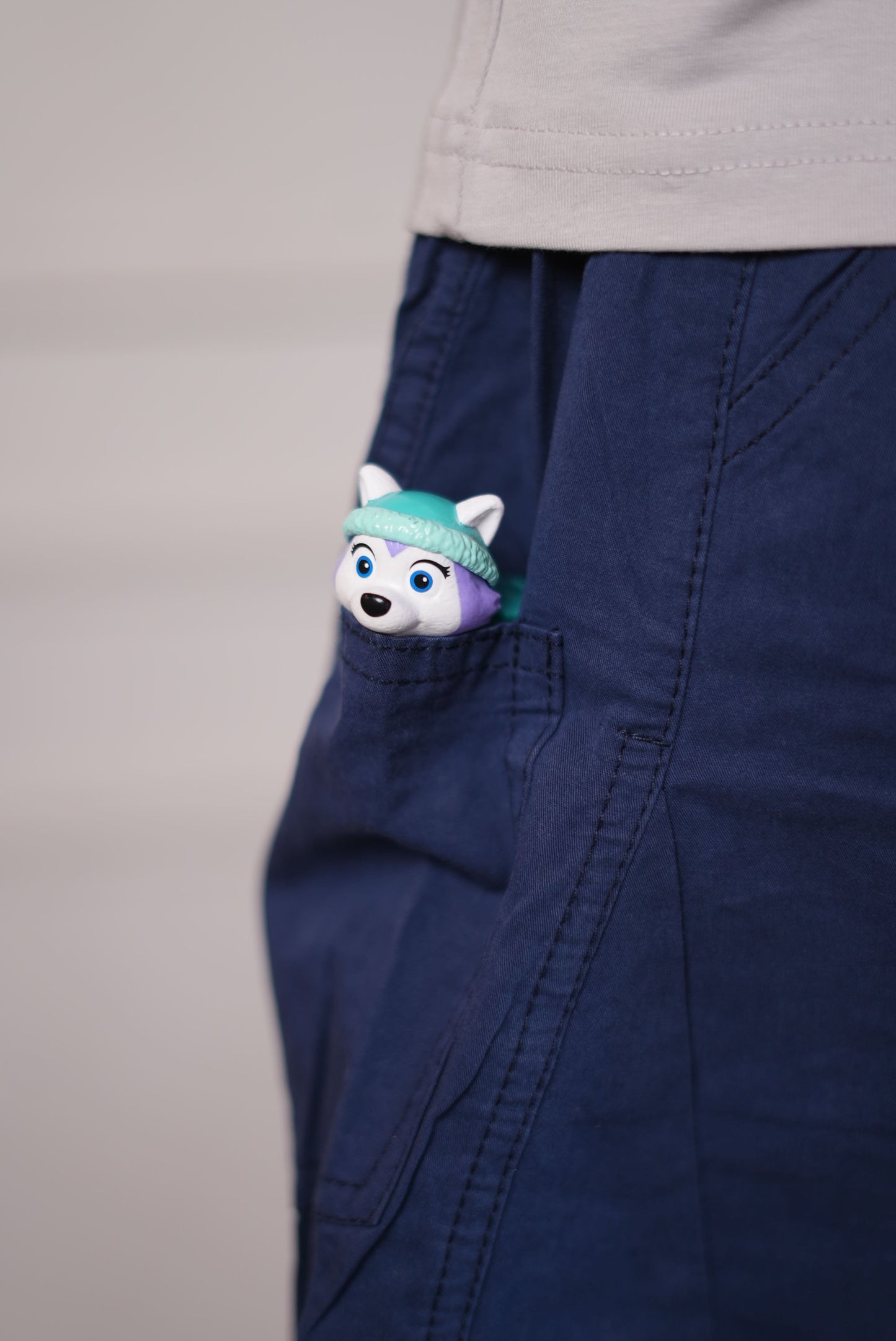 Small plush toy with a teal ribbon peeking out from a pocket of blue pants on a neutral background