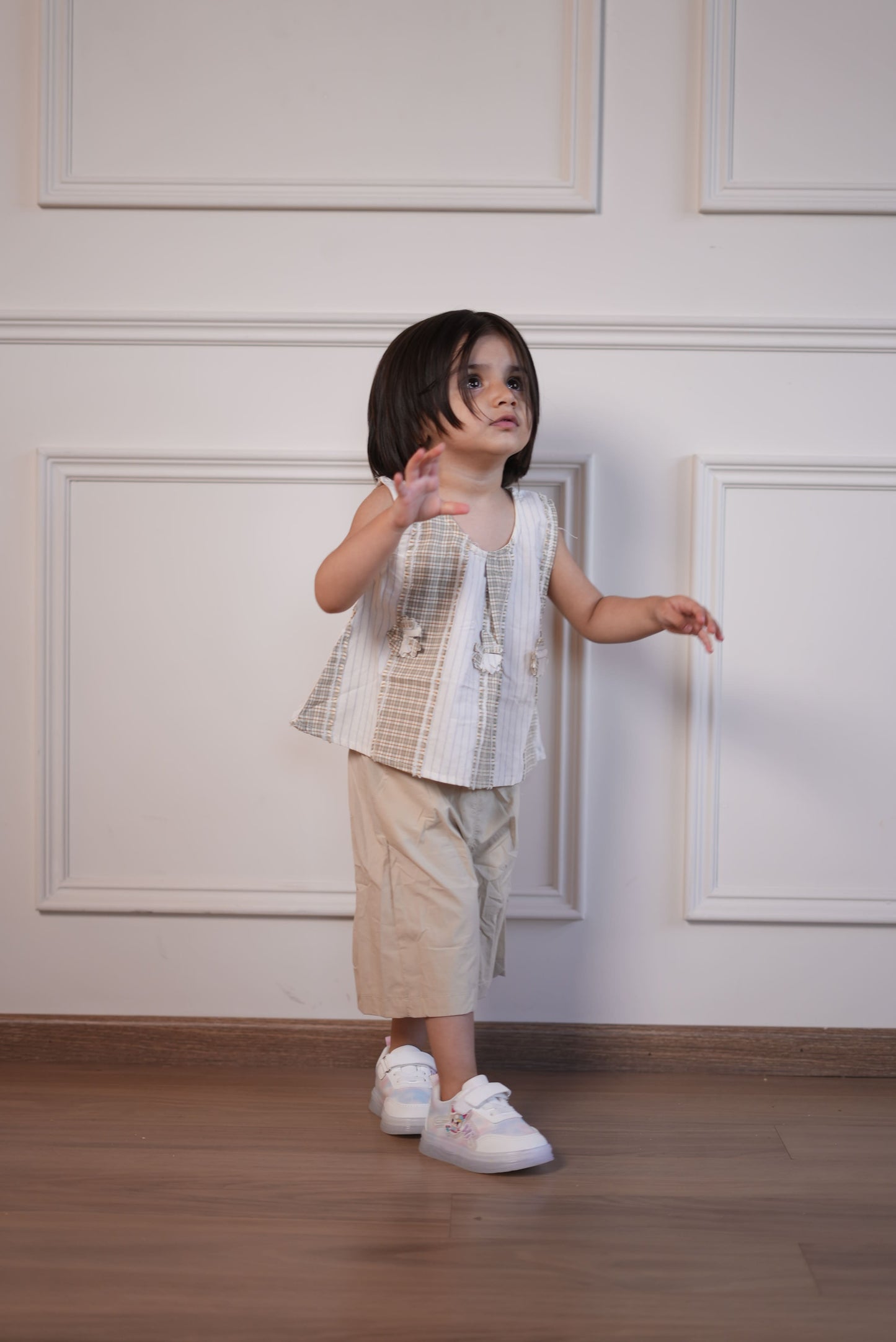Child in a checkered dress standing against a white paneled wall.
