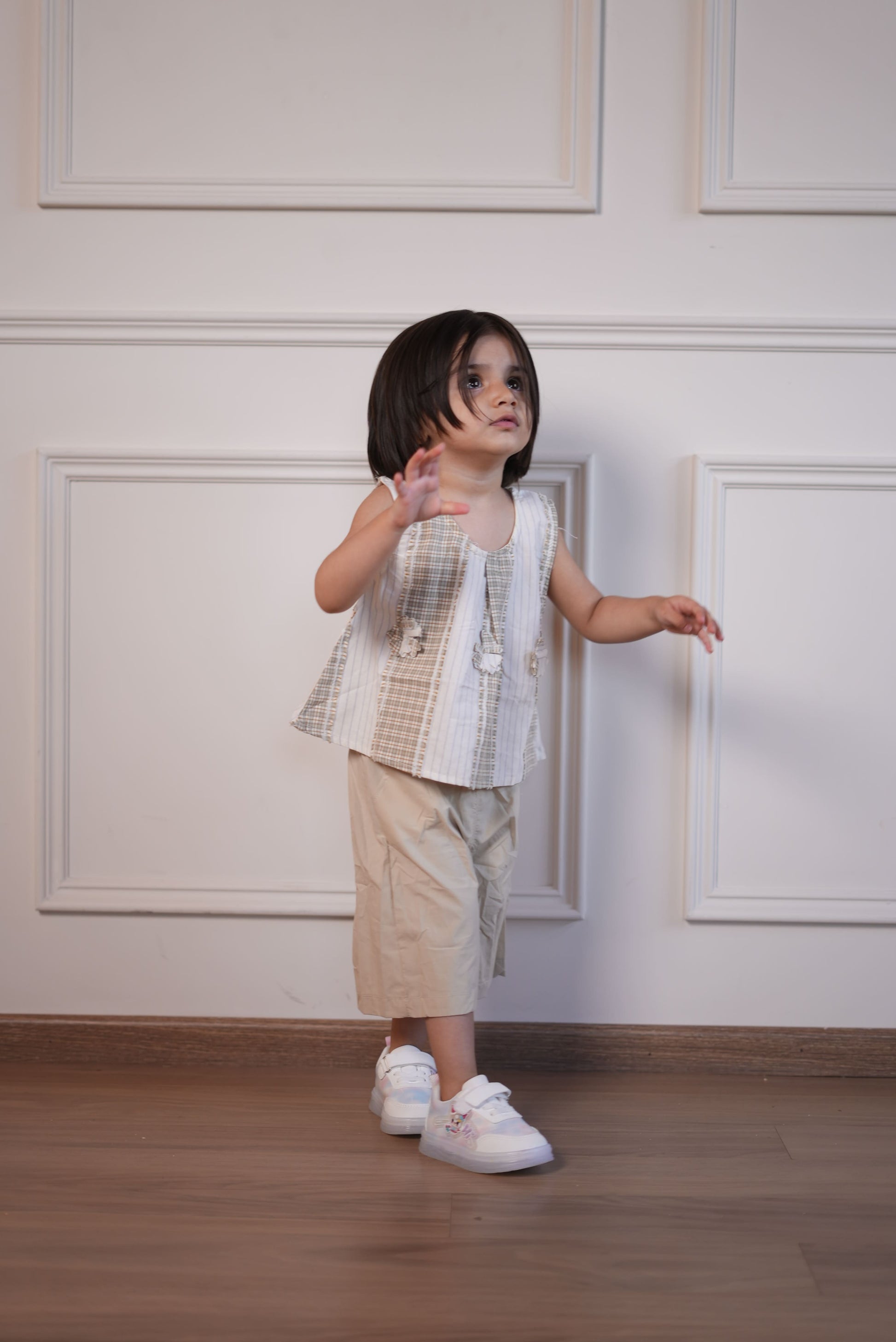 Child in a checkered dress standing against a white paneled wall.