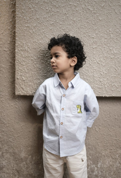 Child wearing a light blue shirt with a small yellow design, standing against a beige wall.