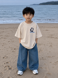 Child standing on a beach wearing a beige t-shirt and blue jeans with a penguin graphic.
