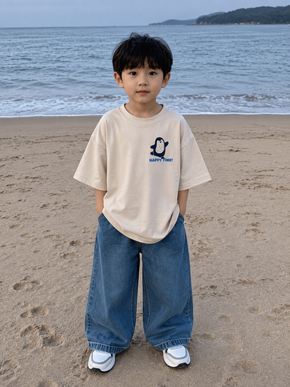Child standing on a beach wearing a beige t-shirt and blue jeans with a penguin graphic.