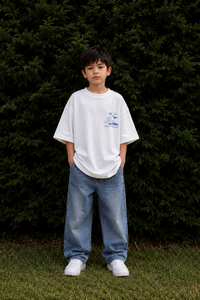 Child wearing a white t-shirt and blue jeans standing in front of dark green bushes.