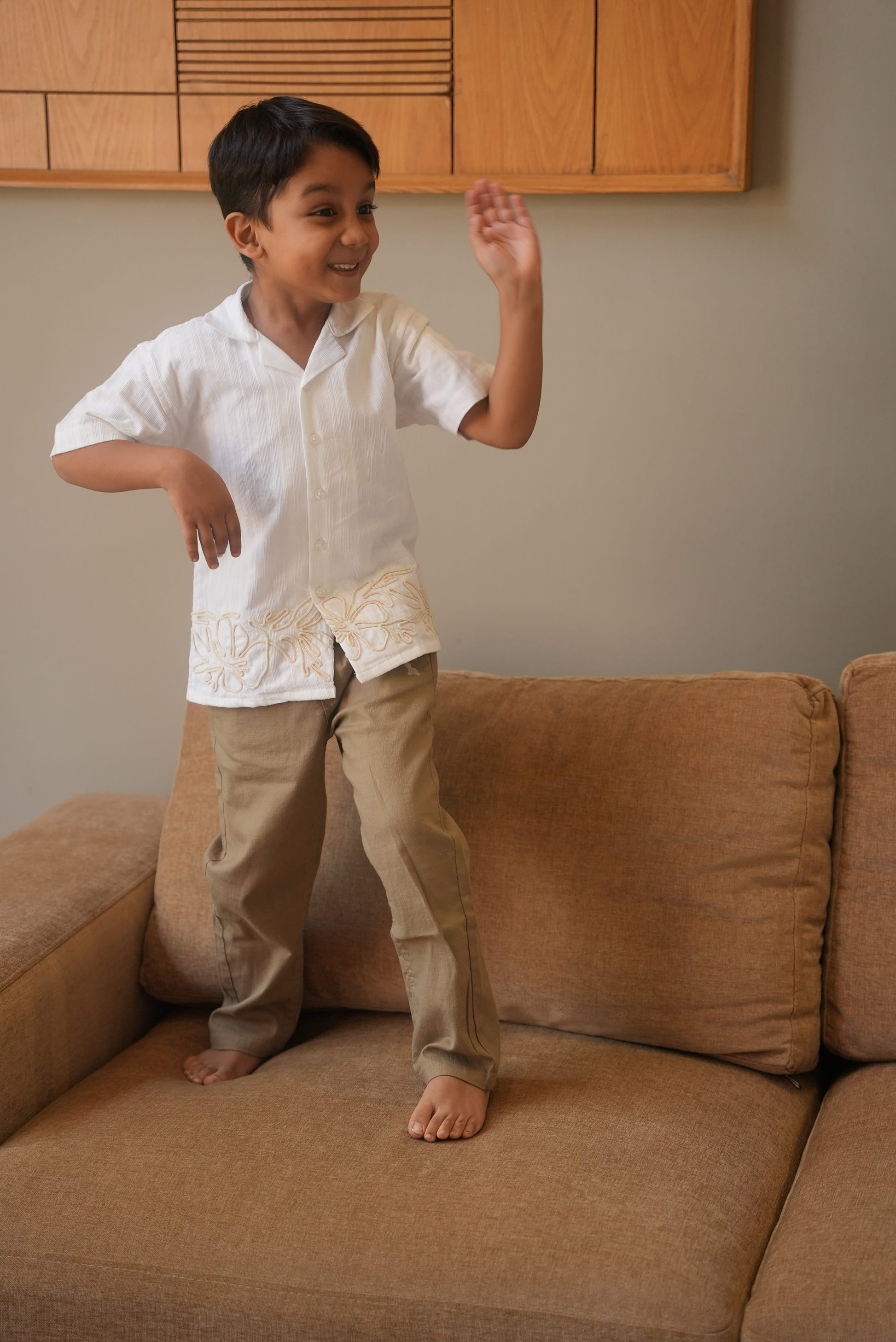 Child standing on a brown couch with a neutral wall and wooden panel in the background