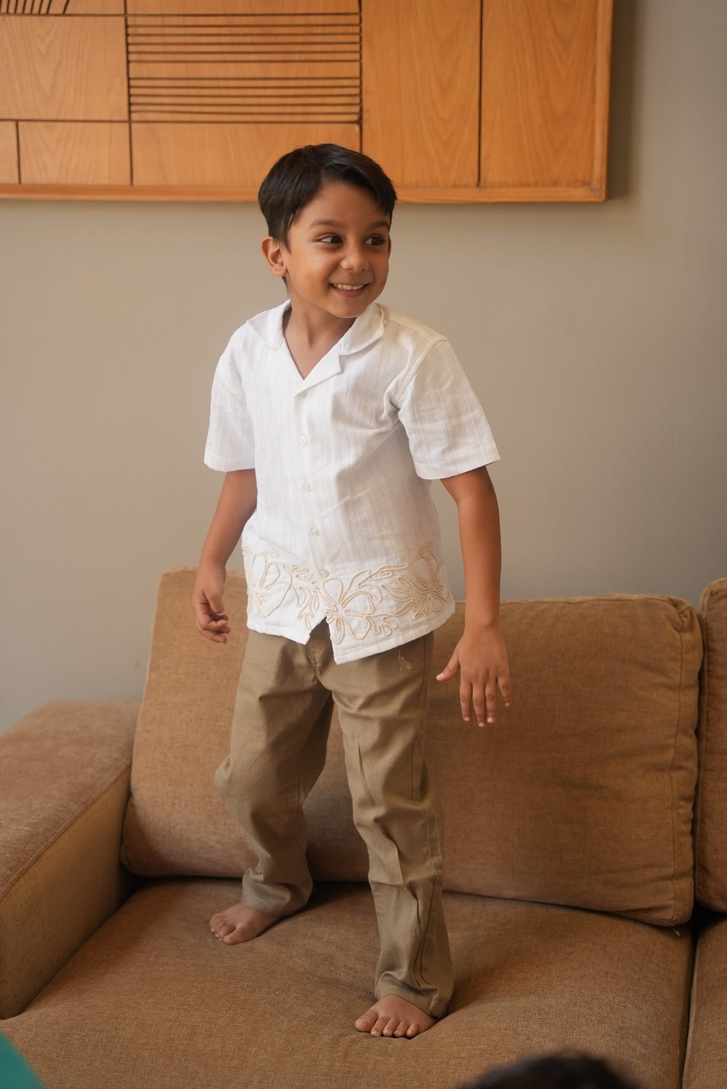 Child in a white shirt and beige pants standing on a brown couch.