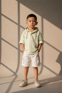Child wearing a light green striped shirt and white shorts standing against a beige wall with shadows.