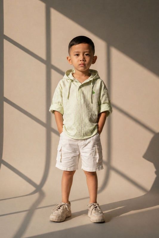Child wearing a light green striped shirt and white shorts standing against a beige wall with shadows.