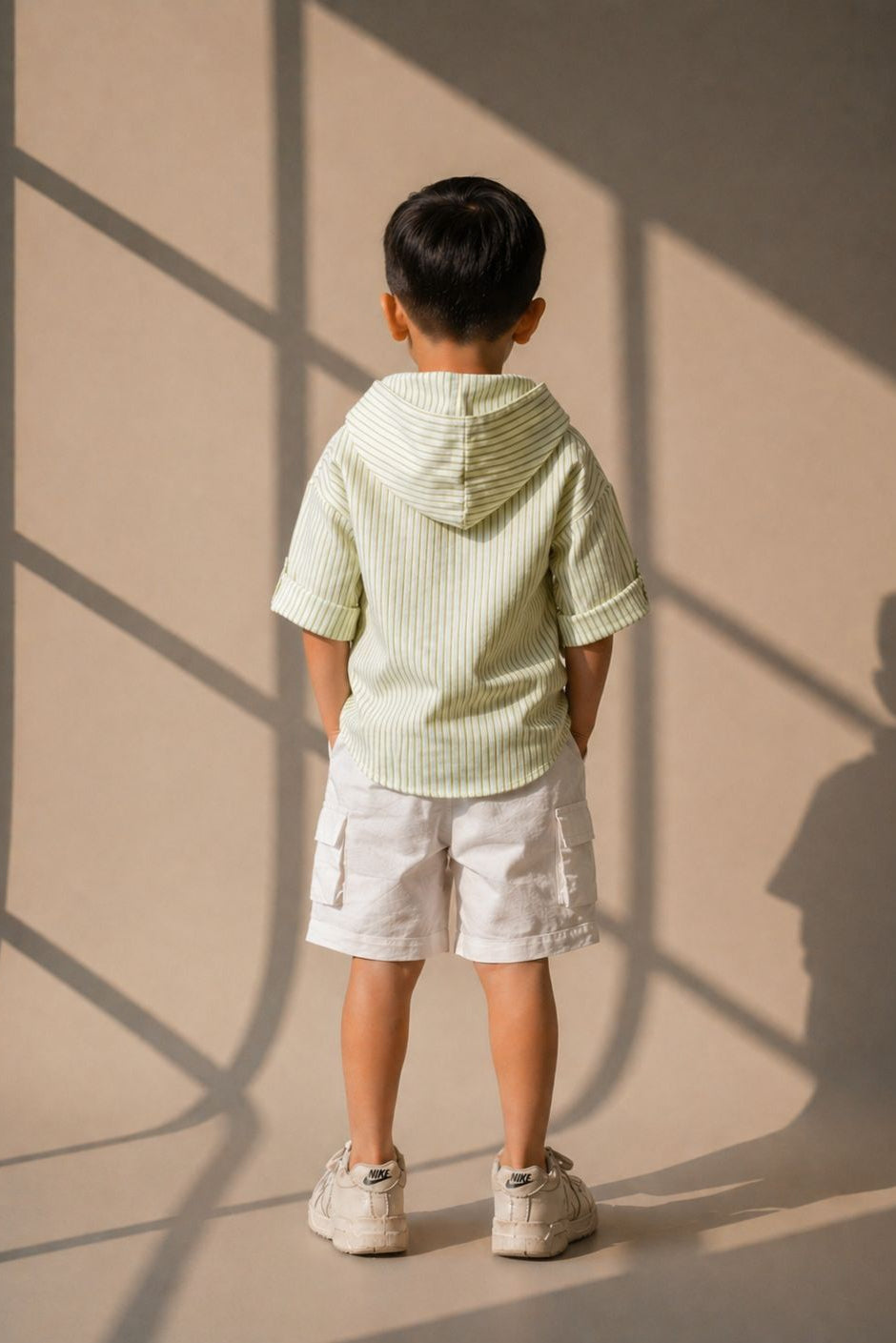 Child wearing a light green striped shirt and white shorts standing against a beige wall with shadows.