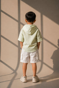 Child wearing a light green striped shirt and white shorts standing against a beige wall with shadows.