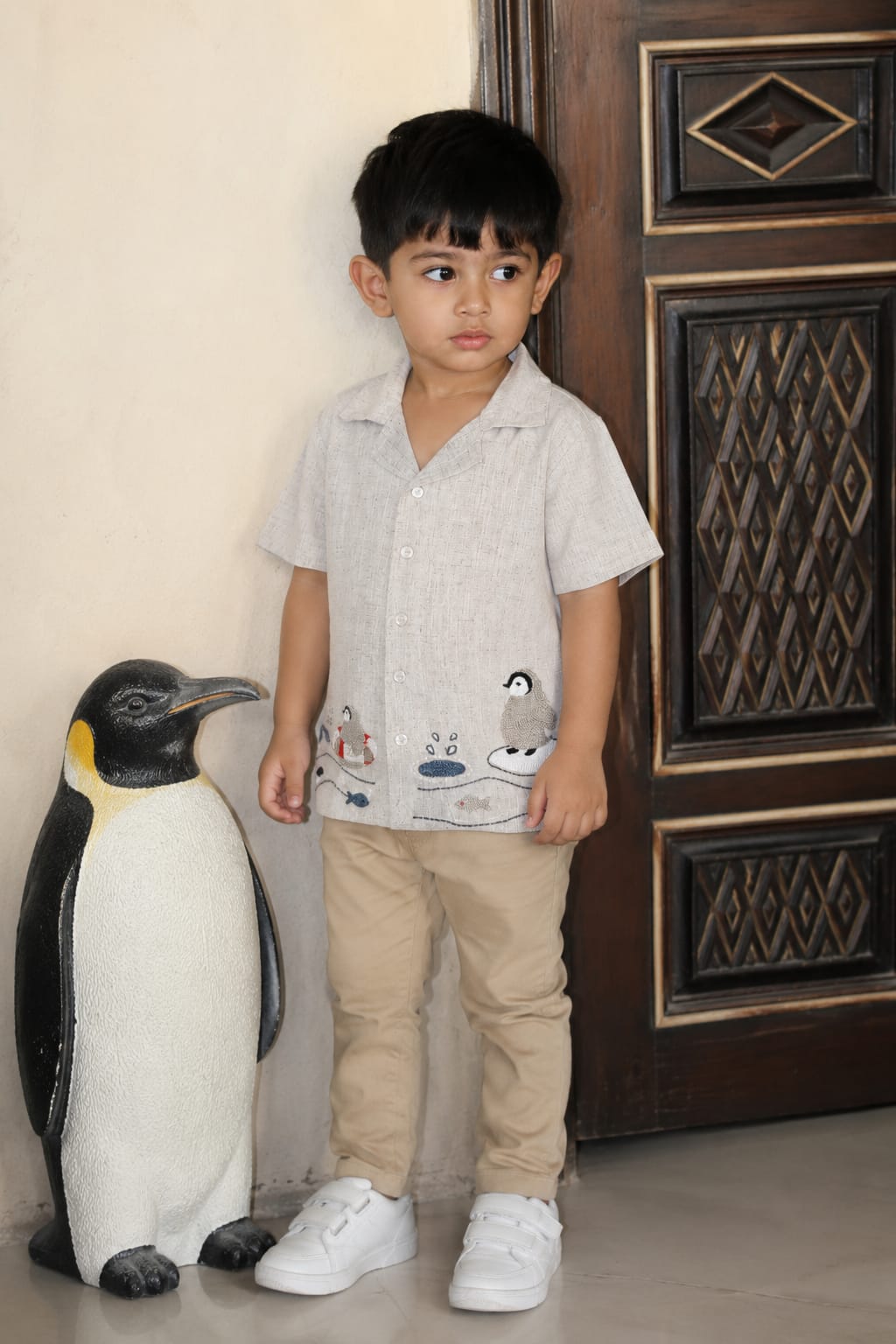 A young boy wearing a light gray short-sleeve shirt with a penguin graphic, beige pants, and white sneakers, standing next to a large penguin toy in front of a dark wooden door.