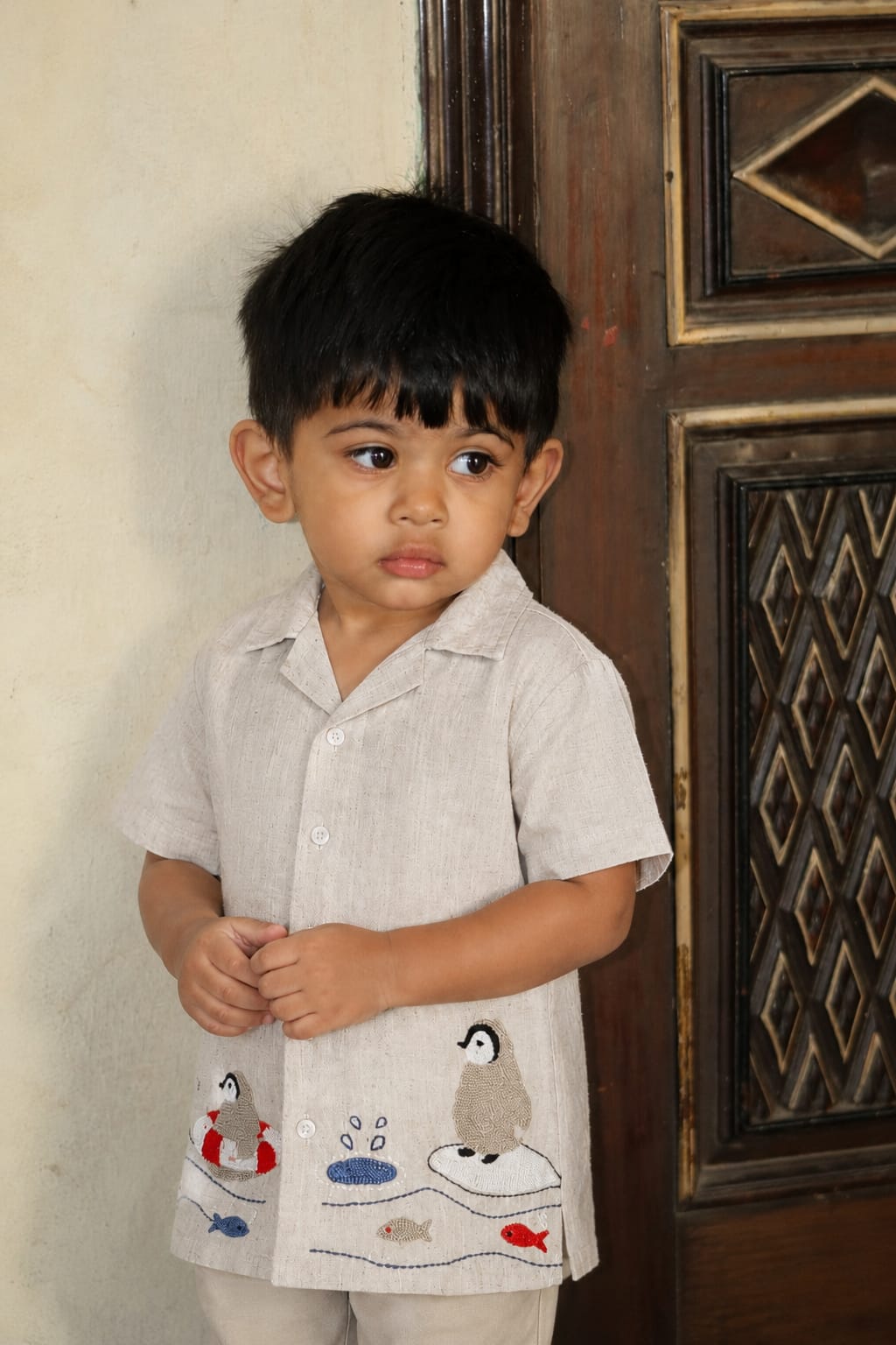 A young boy wearing a light beige short-sleeve button-up shirt with embroidered penguin and fish motifs, standing next to a dark wooden door.