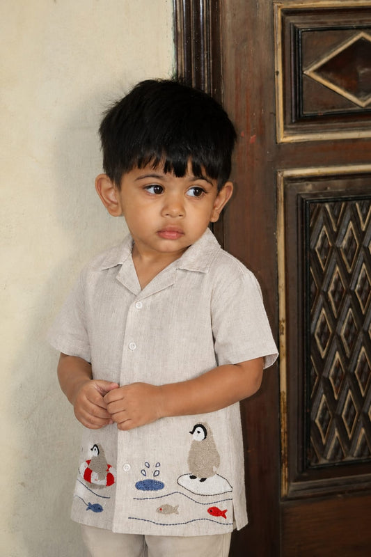 A young boy wearing a light beige short-sleeve button-up shirt with embroidered penguin and fish motifs, standing next to a dark wooden door.