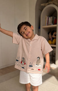 A young boy smiling and posing indoors, wearing a light beige short-sleeve button-up shirt with penguin and fish embroidery, paired with white shorts.