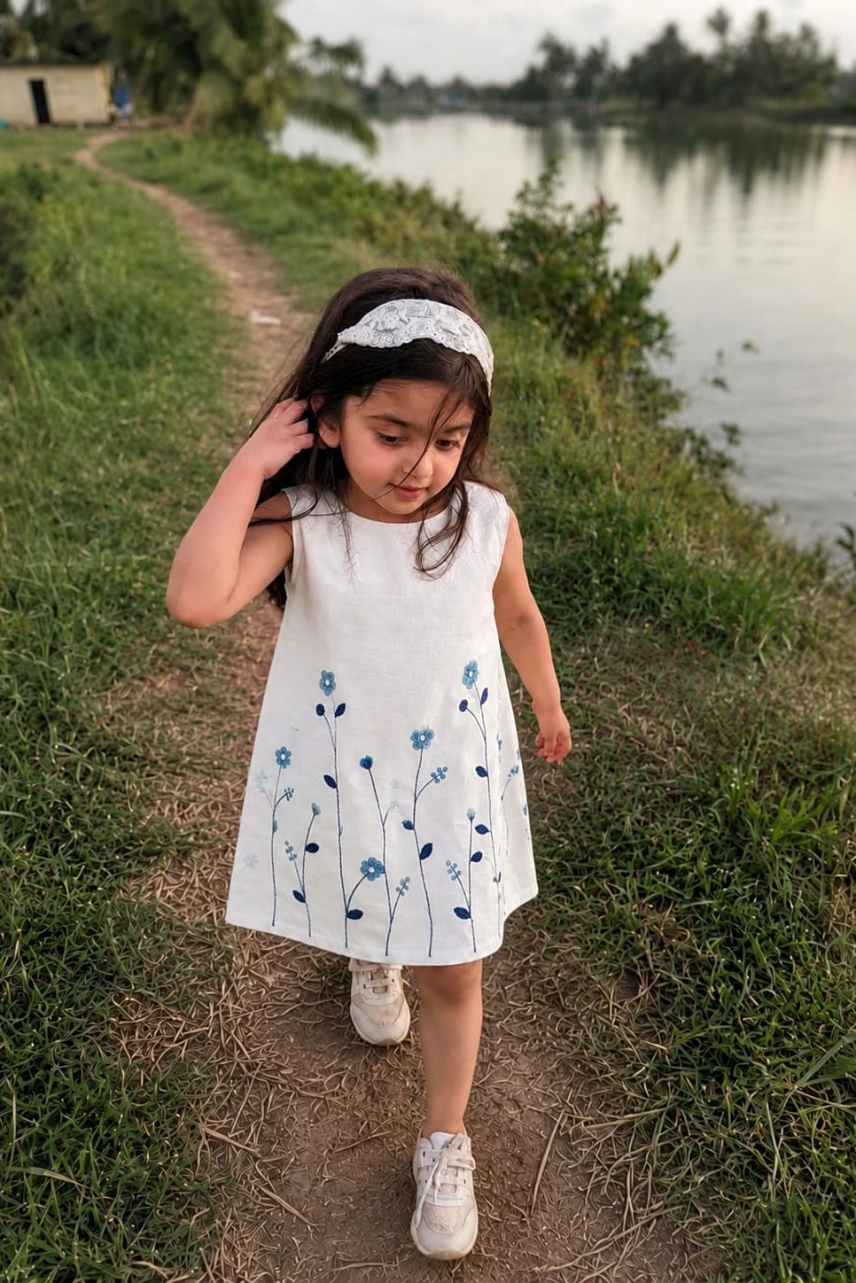 A young girl walking on a dirt path next to a lake, wearing a sleeveless white dress with blue floral embroidery and a white lace headband.