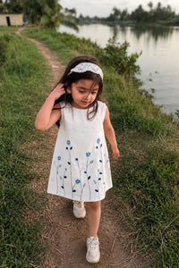 A young girl walking on a dirt path next to a lake, wearing a sleeveless white dress with blue floral embroidery and a white lace headband.