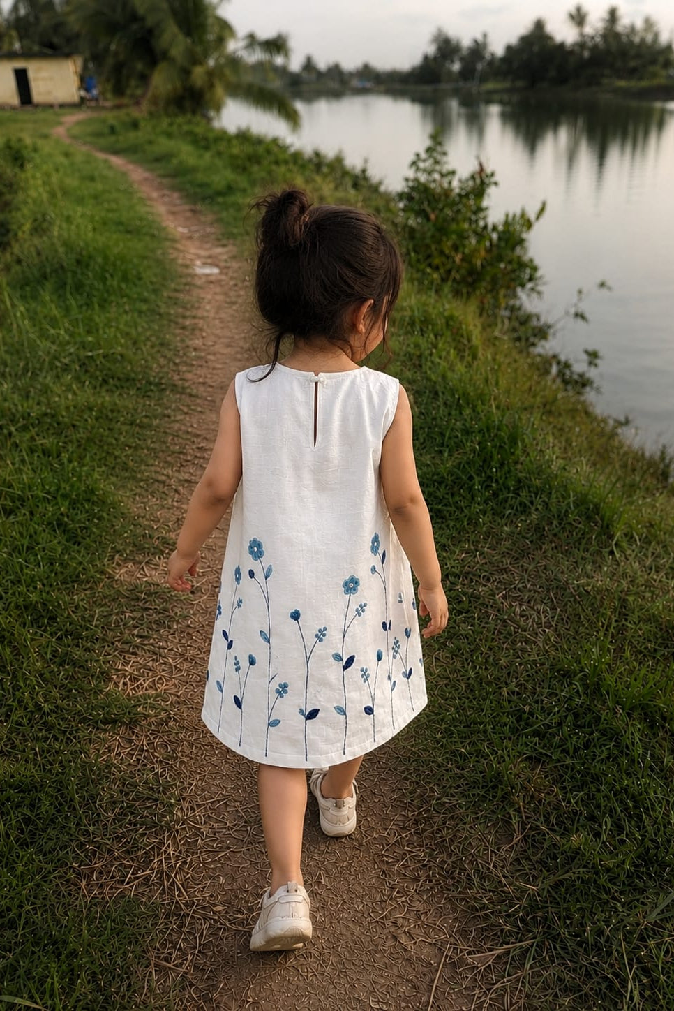 A young child seen from behind walking on a dirt path beside a lake, wearing a sleeveless white dress with blue floral embroidery.